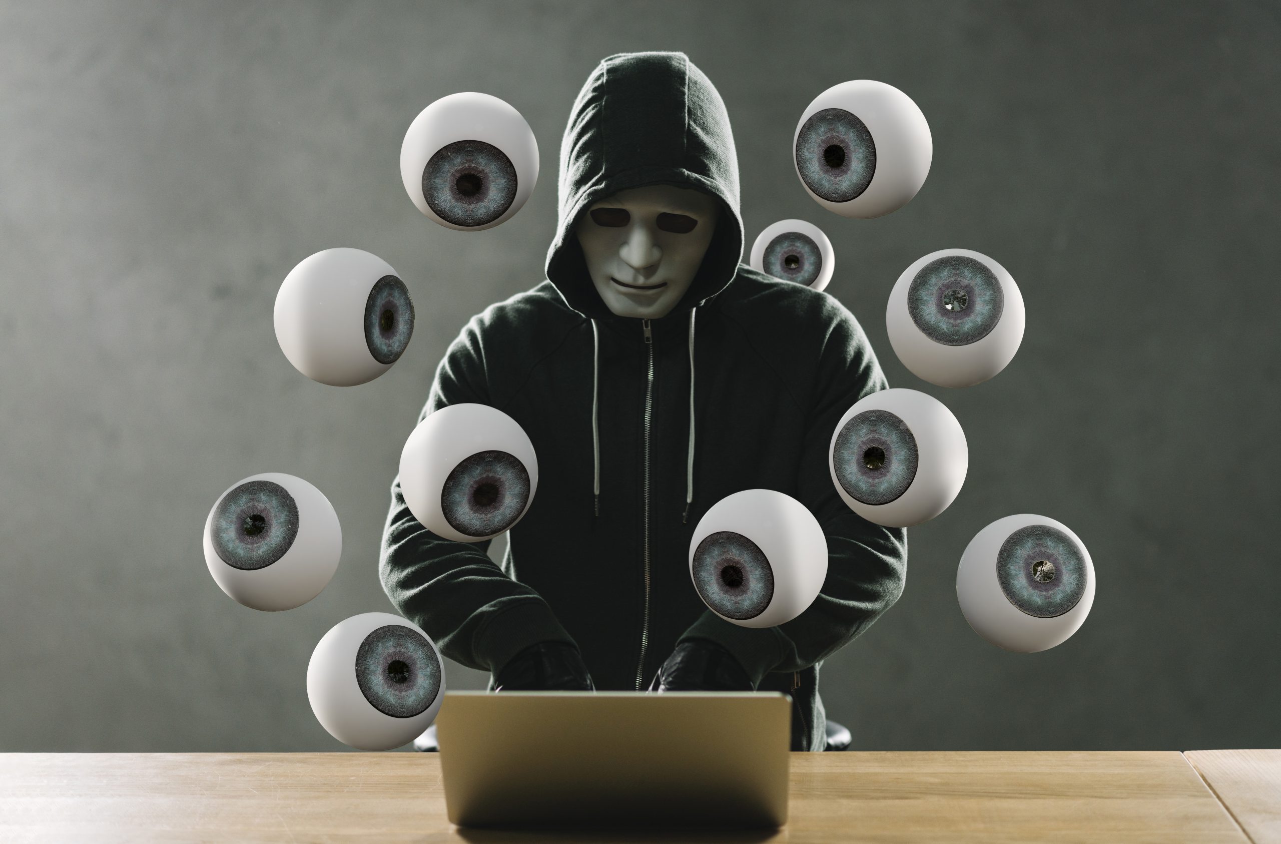 Maskierter Mann tippt auf Laptop wärend viele Augen ihn beobachten.
