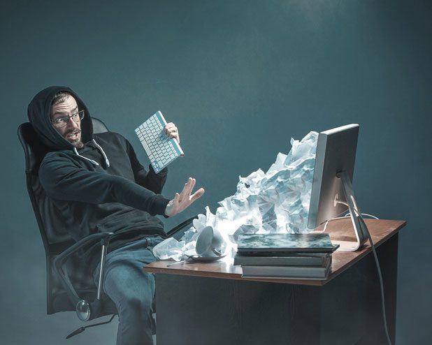 Junger Mann mit Kaputze sitzt erschrocken vor Seinem PC Bildschirm. Aus dem Bildschirm kommen viele Papierkugeln welche den Schreibtisch zumüllen