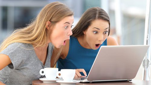Zwei überraschte Frauen stehen mit aufgerissenen Mündern vor ihrem Bildschirm