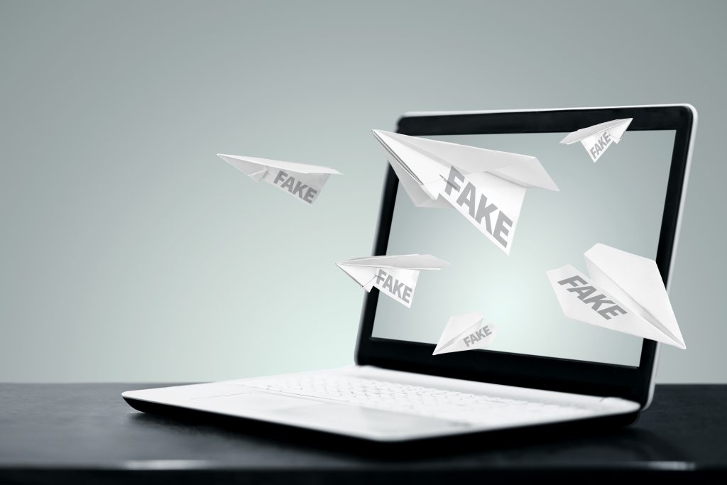 Laptop steht auf einem Tisch. Papierfliger mit der aufsachrift "Fake" fliegen aus dem Bildschirm